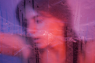 Young attractive long-haired girl in a dress behind transparent plastic foil. Purple lighting. High...