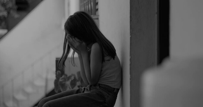 Young child covering face with hands, eyes closed, sitting on the floor in an emotional moment, dealing with trauma and sadness in a quiet, reflective outdoor urban environment, black and white