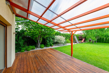 Wooden pergola with transparent polycarbonate roof covering a garden deck in a modern backyard,...