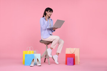 Internet shopping. Happy woman with laptop sitting on stool among colorful bags against pink...