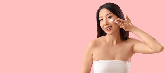 Beautiful Asian woman applying cosmetic cream on her face against pink background with space for...