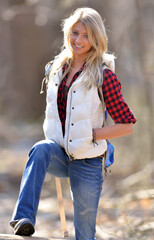 Obraz premium Stunning young blonde model in a a red and black flannel shirt, white down vest, and jeans on hike in woods - climbing over log