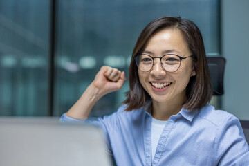 Successful Asian woman close up at workplace inside office. Businesswoman using laptop at work, received online notification about successful results and achievements, celebrating triumph.