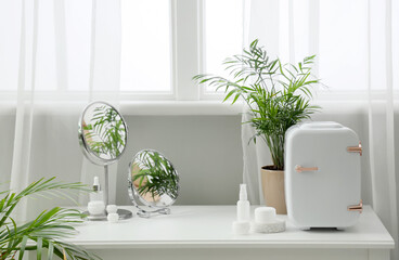 Table with small refrigerator, houseplant in pot, mirrors and cosmetic products near window