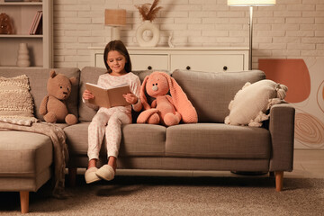 Cute little girl reading book while sitting on sofa with toys in living room