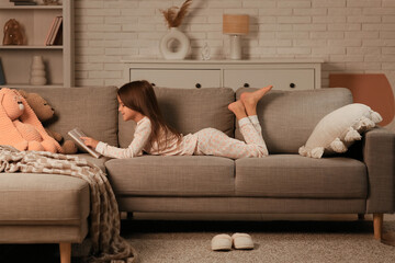 Cute little girl reading book while lying on sofa in living room