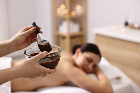 Spa worker with cosmetic product for chocolate body wrap near woman in salon, closeup. Selective focus