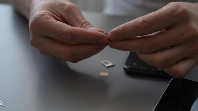 Technician inserting sim card into smartphone, silver ejector pin and tray resting on grey workspace, showcasing precise mobile device maintenance. Process of changing mobile service providers