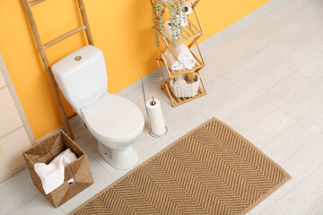 Interior of restroom with toilet bowl, basket with towel and shelving unit near yellow wall