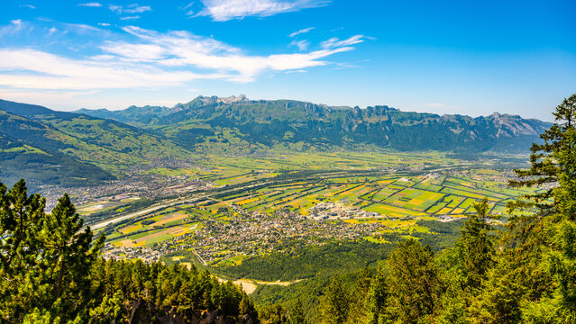 A stunning vista from Gaflei lookout tower showcases the picturesque town of Schaan, surrounded by lush greenery and mountain ranges under a clear blue sky.