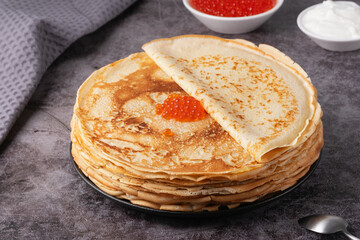 Freshly made pancakes stacked high with red caviar on top and sour cream in bowls nearby
