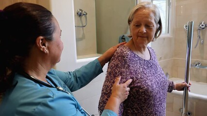 A caregiver offers support to an elderly woman in a bathroom, ensuring her safety while preparing for a bath. Both appear attentive and focused on the task