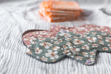 Reusable cloth menstrual pads with floral patterns in pink and green tones placed on a textured white fabric, with disposable pads in the background, promoting sustainable feminine care.