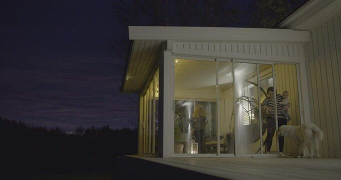 Mother opening sliding door at night to let a dog inside while family is gathered indoors