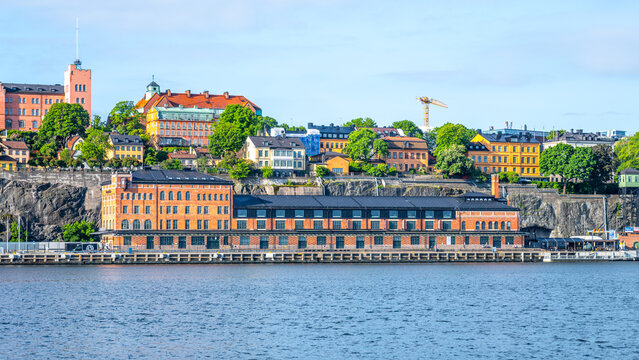 The waterfront view of Fotografiska museum in Stockholm, Sweden, basks under a clear blue sky, with vibrant buildings atop the rocky terrain in the background. - Powered by Adobe