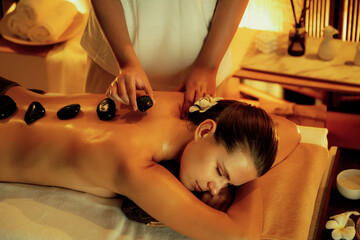 Fototapeta premium Hot stone massage at spa salon in luxury resort with warm candle light, blissful woman customer enjoying spa basalt stone massage glide over body with soothing warmth. Quiescent