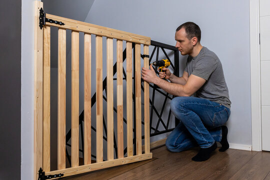 A man kneels while using a yellow and black power drill to install a wooden safety gate with black metal hinges near a staircase with geometric railing.