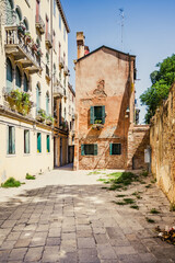 A peaceful alleyway in Venice showcases beautiful architecture with a weathered building and vibrant greens. The sun illuminates the street, highlighting its old-world charm and quaint atmosphere.