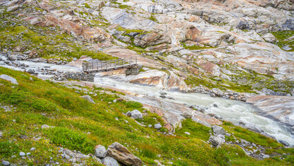 A wooden bridge spans a rushing stream sourced from the Schlaten Glacier, surrounded by rocky terrain and vibrant green vegetation in Hohe Tauern National Park.