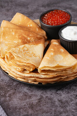 Freshly made pancakes stacked high with red caviar on top and sour cream in bowls nearby