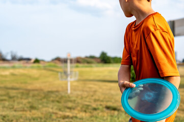 Selective focus fairway grass with a defocused young child holding a putter disc and golf goal in...