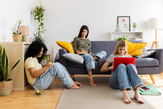 Three female students studying together at home, sitting comfortably on the floor and sofa, engaged in their academic assignments. Female friendship and youth concept