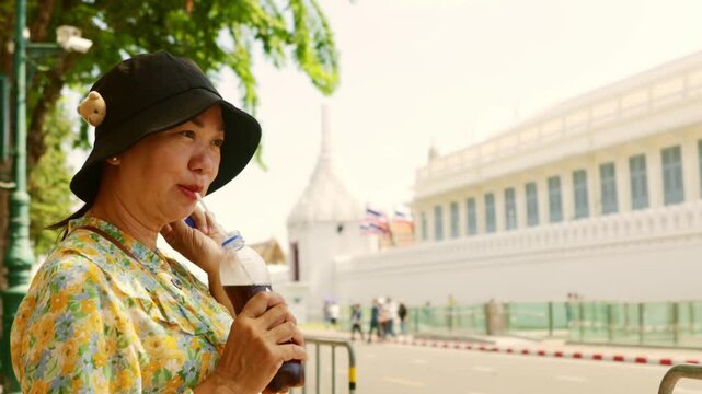 Asian adult woman tourist happily visiting Wat Phra Kaew and Grand Palace in Bangkok, Thailand taking break from the sweltering heat under tree with refreshing soda drink.