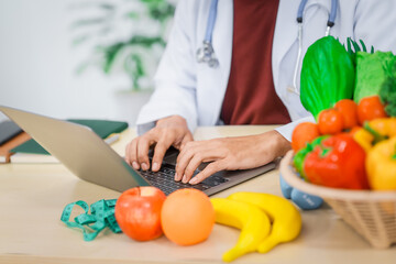 An Asian male nutritionist with a beard, wearing a lab coat, sits at a desk for an online consultation. His workspace includes fruits, vegetables, laptop, health supplements, promoting wellness.