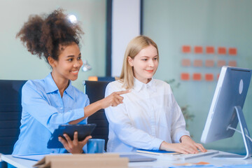 Obraz premium Young African American woman and Russian woman work together in business office, consulting on financial charts using desktop PC and tablet. They discuss strategies and collaborate as colleagues.