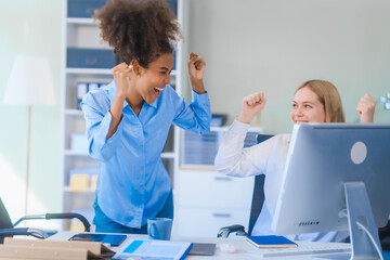 Young African American woman and Russian woman work together in business office, consulting on financial charts using desktop PC and tablet. They discuss strategies and collaborate as colleagues.