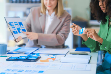 A young African American woman and a young Russian woman work together in a business office. They discuss UX design, using color swatches and tablets, while planning layouts and interfaces.