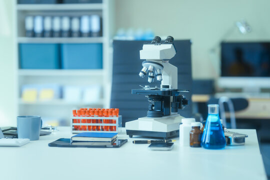 In an empty lab room with no people, a desk holds a blood tube, virus samples, and a monitor displaying blood cell analysis. A medical lab technician works with advanced equipment.