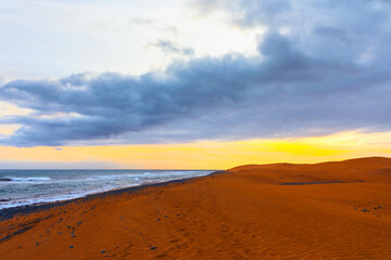 Coastal landscape with a sandy beach extending into distance, bordered by ocean on left and sand dunes on right. Sky is partly cloudy with mix of dark and light clouds, peaceful and picturesque scene