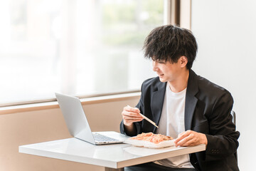イートインスペース・社員食堂でパソコンを見ながら昼食にお弁当を食べる若いアジア人男性ビジネスマン
