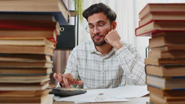 Exhausted Indian businessman reviewing documents reports while using calculator working hard in home office. Stressed alone freelancer touching head in despair surrounded by stacks of books on table.