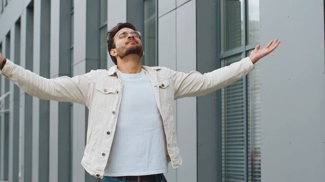 Young Indian man taking a deep breath of fresh air, relaxing, taking a break, resting, meditating, calm down outdoors. Arabian Hindu guy enjoying happy day in downtown city street. Business people
