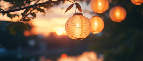 Obraz premium Warm lanterns softly illuminate the evening sky, suspended from branches, as dusk paints the landscape in hues of orange and gold.