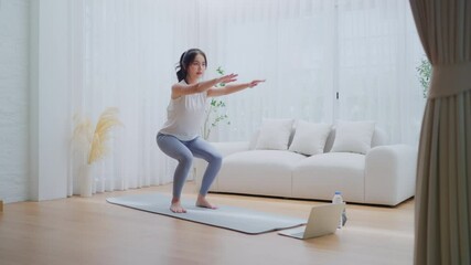 Young Asian beautiful woman doing aerobic exercise at home. Attractive girl doing  workout by squatting, follow instructions video from online trainer for health in house