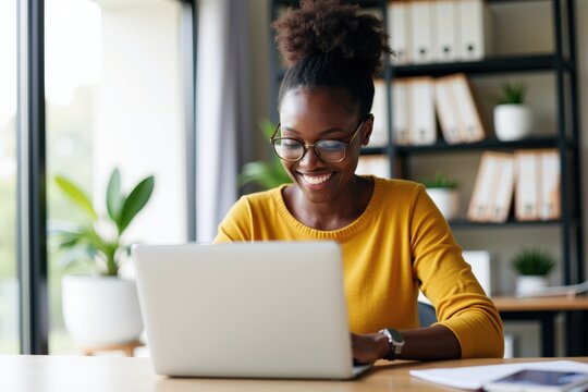 Happy Young Woman Working on Laptop in Bright Modern Office with Plants and Books, Creating Content or Engaging in Remote Work