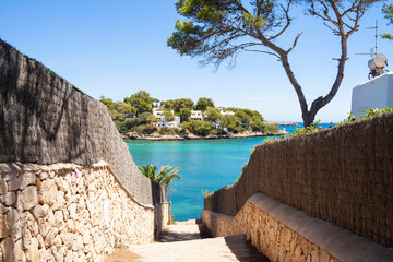 A stone pathway leads to a turquoise bay, framed by lush greenery and white houses. The clear blue...