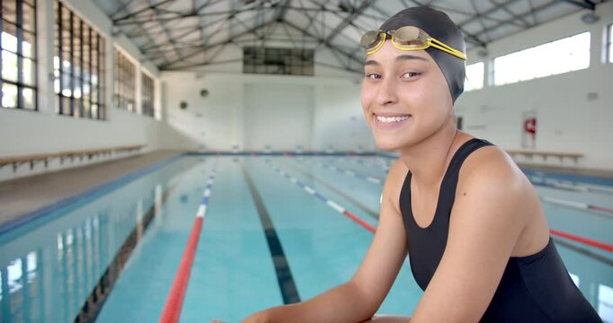 Smiling Female Swimmer in swim cap and goggles sitting by indoor pool, copy space