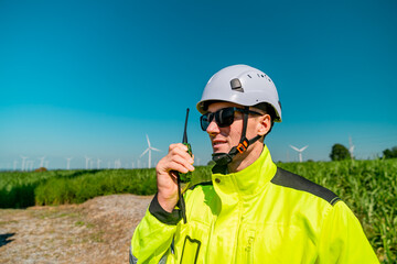 Worker in bright gear communicates with walkie-talkie in a field with wind turbines under clear...