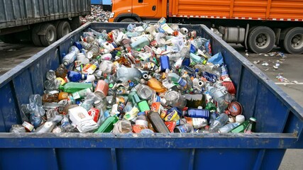 A blue bin full of plastic waste is filled with various types and colors, such as bottles, packaging materials, glassware, paper, and aluminum cans.