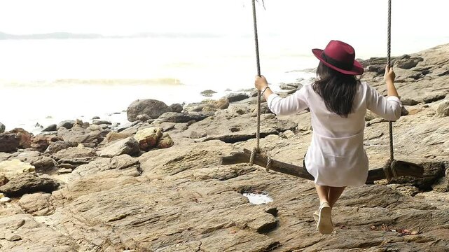 Serene woman in a red hat swings on a tree rope overlooking rocky terrain by the sea, surrounded by lush greenery and nature's tranquility