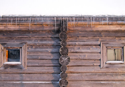 Wooden log cabin wall with two small windows and icicles hanging from the snowy roof, showing traditional winter architecture.