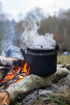 A black kettle sitting on a smoldering log over an open campfire, with steam rising from its spout and lid.
