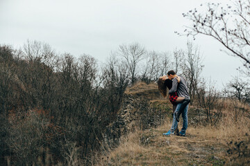 a couple standing in an open area, perhaps on a hill, with dry grass and golden trees around them, which could be seen in autumn or winter. The sky is cloudy.