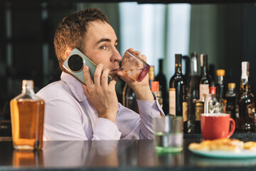 An drunk businessman talking on phone while holding a glass of alcohol in his hands at a bar in the evening. Problems at work and alcoholism. A man in a shirt drinks rum and talks on the phone.