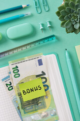 Euro banknotes with a sticker with the word Bonus in English lying on an open notebook. Mint-colored stationery is in the background.
