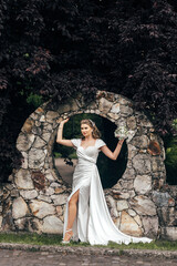 bride sitting against the background of a stone wall with a round hole. She is holding a small...